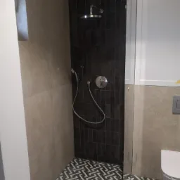 Nowoczesna kabina prysznicowa z czarnymi, wąskimi płytkami, deszczownicą i baterią termostatyczną, uzupełniona geometryczną mozaiką podłogową i beżowymi płytami ściennymi z wnęką.