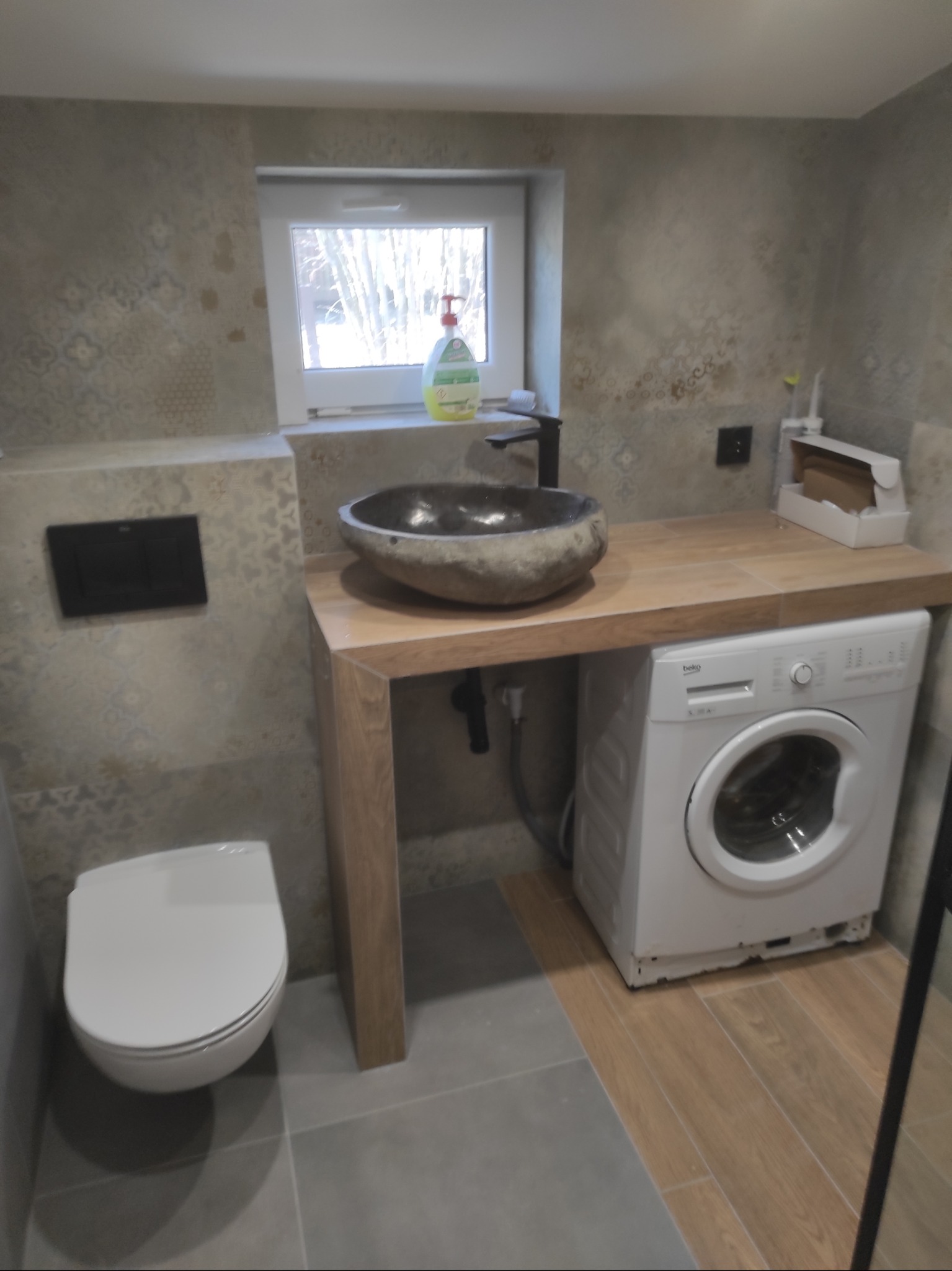 Nowoczesna łazienka z umywalką w kształcie kamienia, drewnianym blatem, pralką i wiszącą toaletą, wykończona płytkami w odcieniach szarości i beżu.