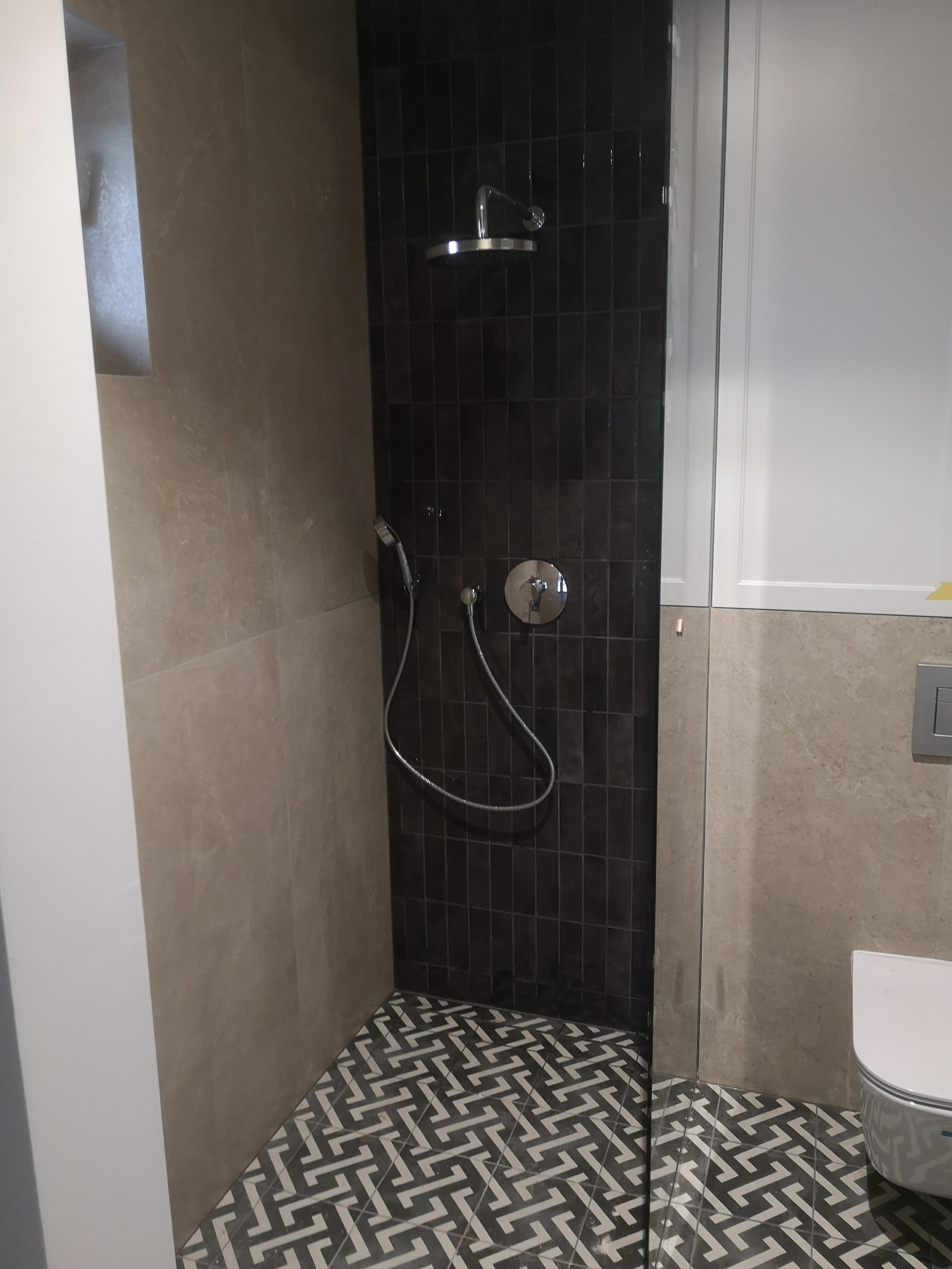 Nowoczesna kabina prysznicowa z czarnymi, wąskimi płytkami, deszczownicą i baterią termostatyczną, uzupełniona geometryczną mozaiką podłogową i beżowymi płytami ściennymi z wnęką.