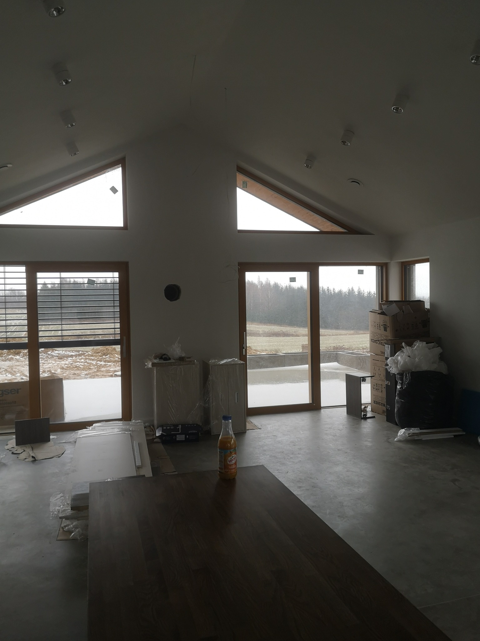 Wnętrze domu w trakcie prac wykończeniowych: widoczne okna, drzwi balkonowe, kartony, butelka soku i fragment blatu stołu na surowym betonowym podłożu.