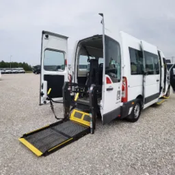 Biały bus z otwartymi drzwiami i rozłożoną platformą dla osób niepełnosprawnych na zewnątrz, na parkingu z szarym żwirem. Widoczny znak osoby na wózku inwalidzkim.
