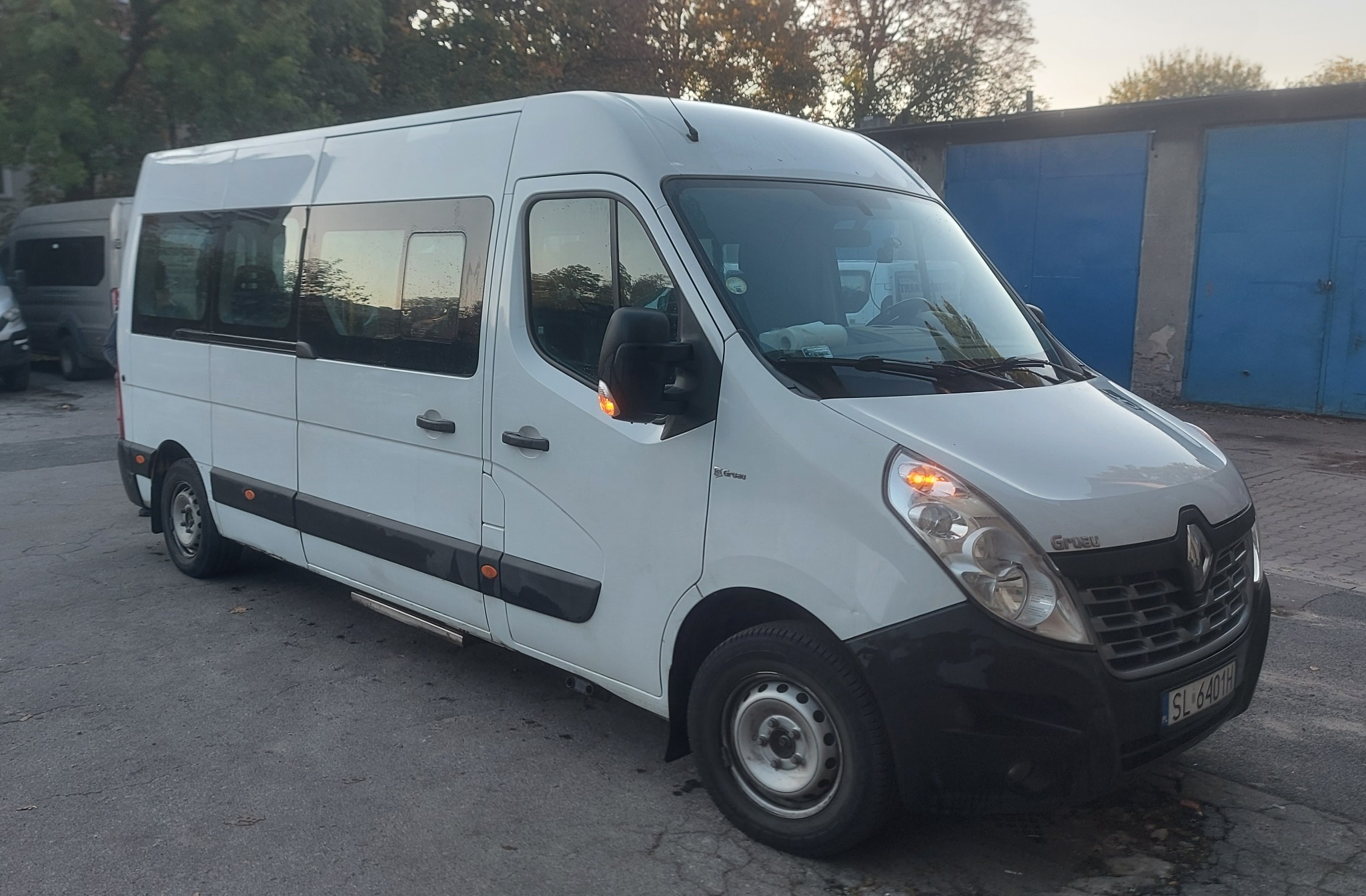 Biały bus Renault Master z włączonymi światłami pozycyjnymi, zaparkowany na tle garażu z niebieskimi drzwiami.