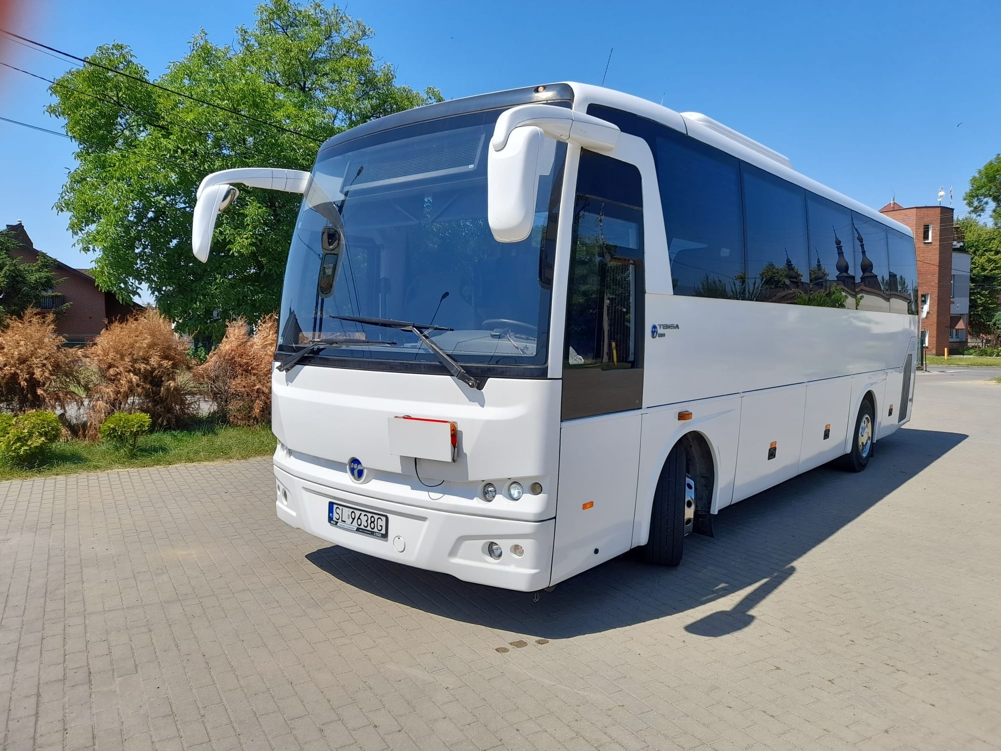 Biały autokar Temsa 120 na kostce brukowej w słoneczny dzień, z odbiciami budynków w oknach.