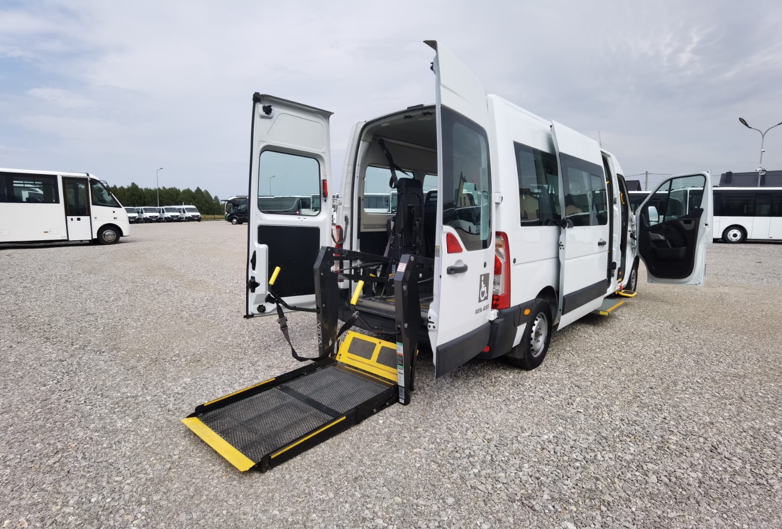 Biały bus z otwartymi drzwiami i rozłożoną platformą dla osób niepełnosprawnych na zewnątrz, na parkingu z szarym żwirem. Widoczny znak osoby na wózku inwalidzkim.