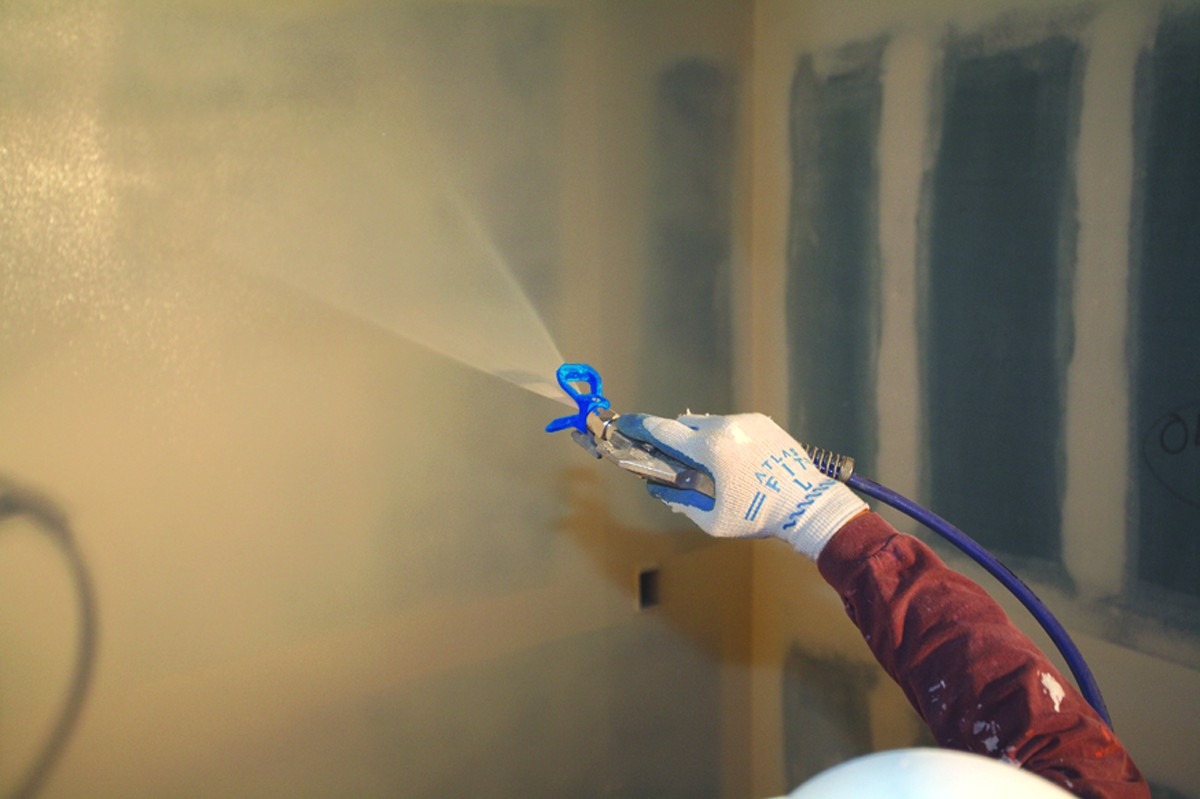 Natryskowe nakładanie gładzi na ścianę z użyciem pistoletu, widoczna ręka w rękawicy ochronnej i niebieski przewód.