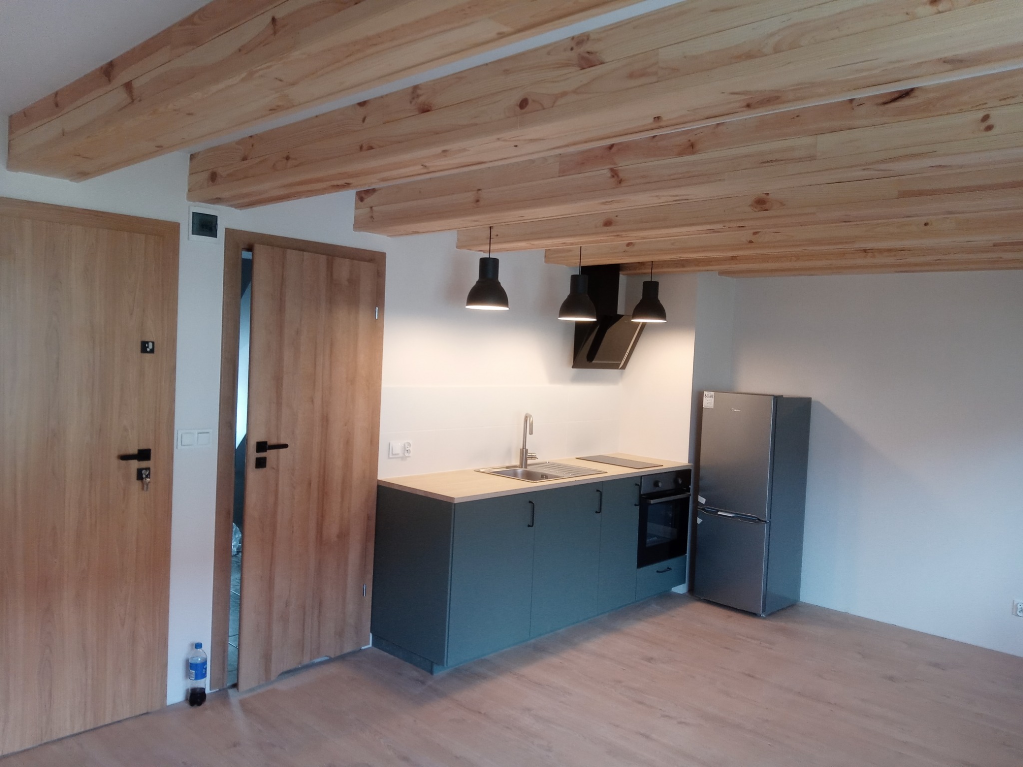 Nowoczesna kuchnia z drewnianym sufitem belkowym, szarymi szafkami, srebrną lodówką i trzema czarnymi lampami wiszącymi, z widoczną butelką wody przy drzwiach.