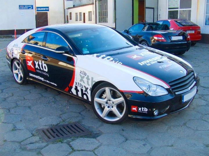 Mercedes CLS z okleiną reklamową XTB, zaparkowany na zewnątrz. Widoczne logo i hasła inwestycyjne. W tle inne samochody i fragment budynku.