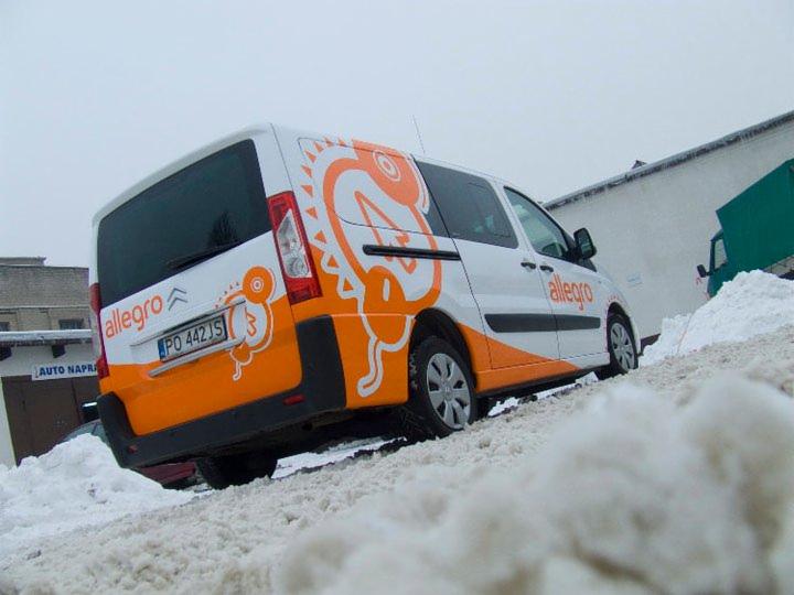 Biały samochód dostawczy z pomarańczowym logo Allegro na śniegu, widok z tyłu pod kątem. Grafika na pojeździe z motywem smoka. Zima.