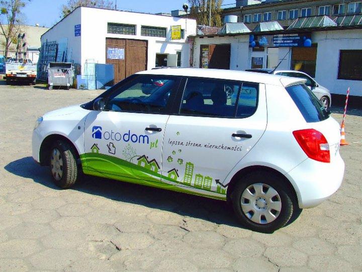 Biała Skoda Fabia z grafiką OtoDom.pl, prezentująca identyfikację wizualną firmy. Zielona linia z domami i napis 'lepsza strona nieruchomości'.