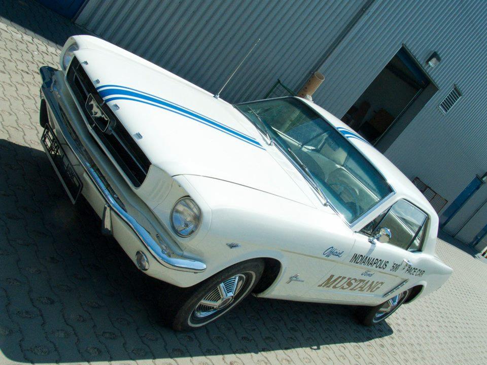 Biały Ford Mustang Indianapolis 500 Pace Car z niebieskimi pasami, zaparkowany na zewnątrz budynku z metalowymi panelami i otwartymi drzwiami garażowymi.