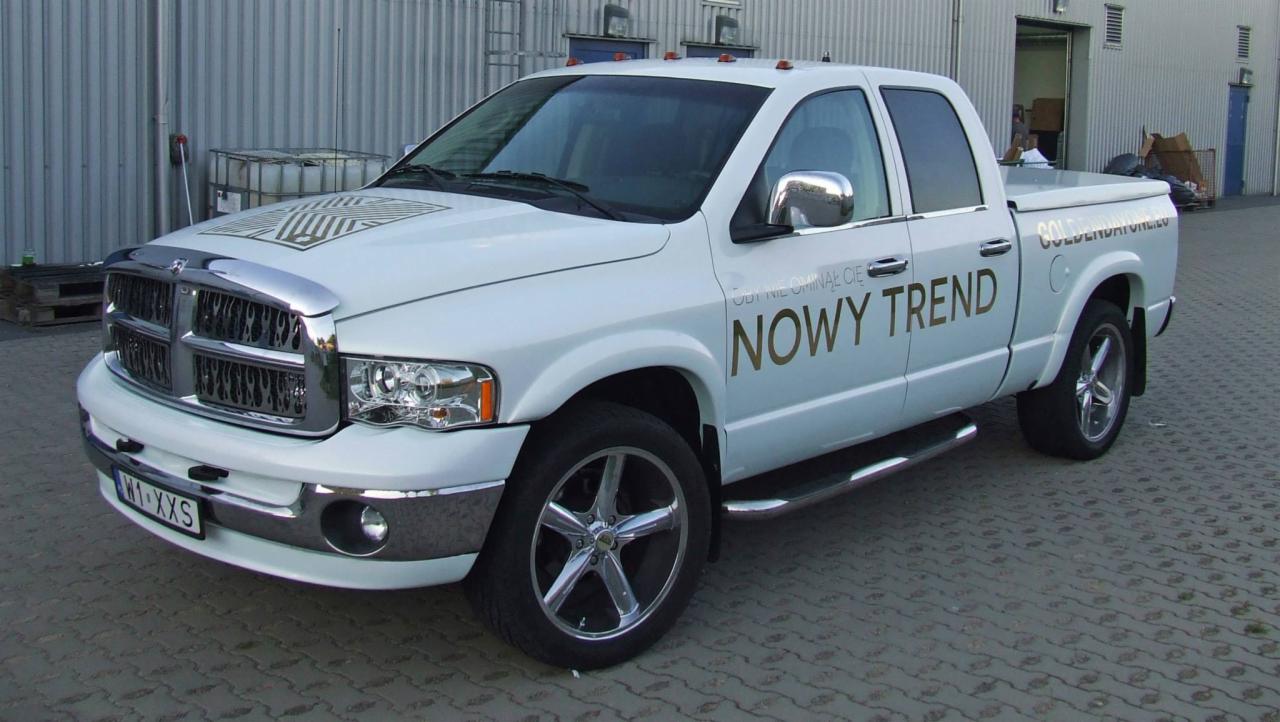 Biały Dodge Ram z złotym napisem 'NOWY TREND' na drzwiach, prezentujący identyfikację wizualną firmy. Widoczny wzór graficzny na masce i logo na tylnej klapie.