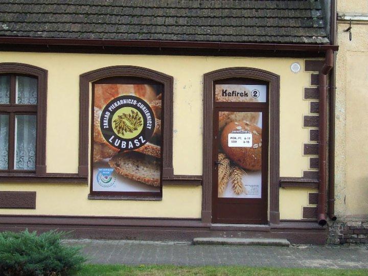 Elewacja piekarni 'Kefirek 2' w Poznaniu z grafikami w oknach i na drzwiach, prezentującymi pieczywo i logo 'Zakład Piekarniczo-Cukierniczy Lubasz'.