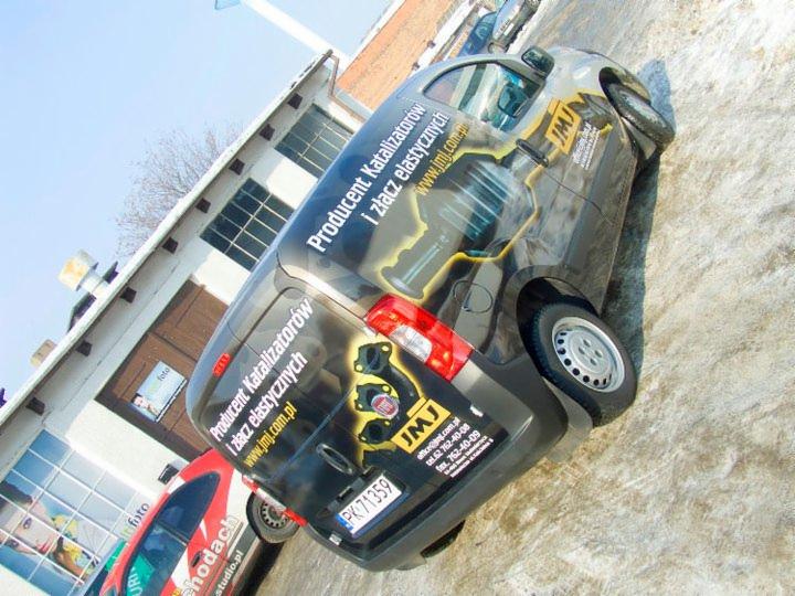 Czarny van z grafiką reklamową JMJ, producenta katalizatorów i złącz elastycznych, zaparkowany na zewnątrz budynku. Widoczne logo firmy i dane kontaktowe.