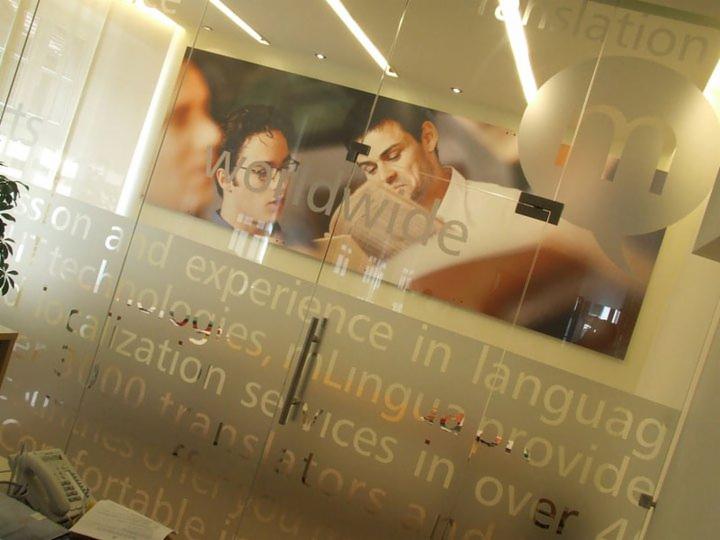 Szklane drzwi biurowe z matową grafiką: logo w dymku, tekst o tłumaczeniach i portret dwóch mężczyzn. Nowoczesne oświetlenie sufitowe.