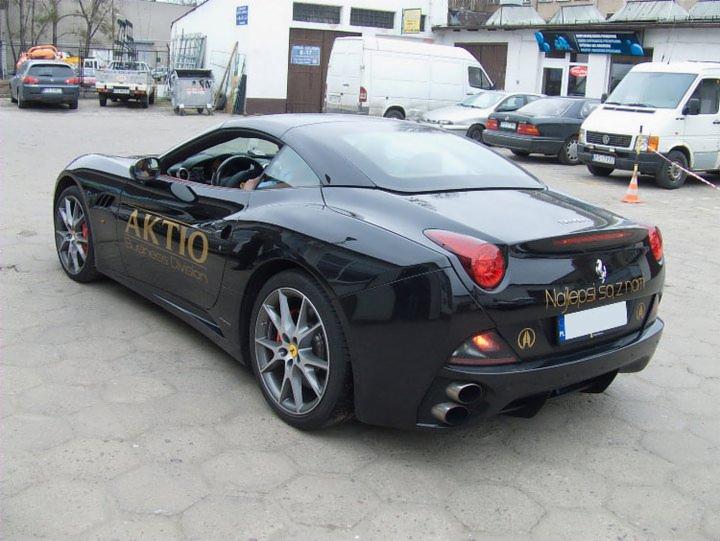 Czarny Ferrari California z logo firmy 'AKTIO' i hasłem 'Najlepsi są z nami' na karoserii, zaparkowany na zewnątrz.