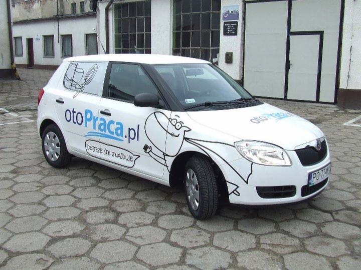Biała Skoda Fabia z reklamą otoPraca.pl, z czarnymi grafikami i niebieskim napisem, zaparkowana na brukowanym podjeździe przed budynkiem.