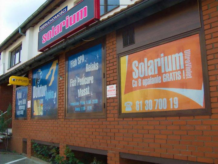 Fasada budynku z szyldami 'Solarium', 'Fish SPA' i 'InPost'. Na ceglanej ścianie okna z reklamami. Widoczny neonowy szyld na dachu.