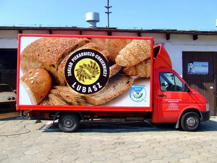 Czerwona furgonetka z grafiką pieczywa i logo piekarni 'Lubasz' na zabudowie, zaparkowana na zewnątrz budynku.