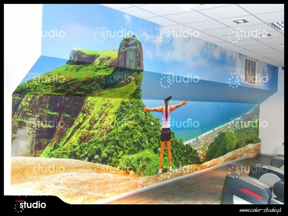 Wnętrze siłowni z fototapetą przedstawiającą górski krajobraz i morze. Kobieta stoi na tle fototapety z rozłożonymi rękami. Widoczne urządzenia do ćwiczeń.