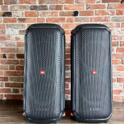 Dwa czarne głośniki estradowe JBL z pomarańczowymi akcentami, ustawione na drewnianej podłodze na tle ceglanej ściany.