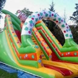 Plenerparty - Kolorowa, dmuchana zjeżdżalnia z motywem małpki i kwiatów, rozłożona na trawie z widocznym niebieskim podkładem zabezpieczającym.