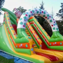 Kolorowa, dmuchana zjeżdżalnia z motywem małpki i kwiatów, rozłożona na trawie z widocznym niebieskim podkładem zabezpieczającym.