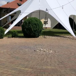 Biały namiot rozłożony na zewnątrz, widoczne okrągłe ułożenie kostki brukowej na ziemi i krzewy w tle, oświetlenie w postaci żarówek zwisających z namiotu.
