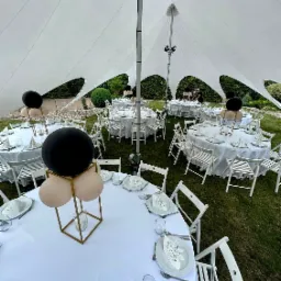 Uroczyste stoły z białymi obrusami i dekoracjami balonowymi w czerni i beżu pod namiotem w kształcie gwiazdy, otoczone zielonym trawnikiem i krzewami.