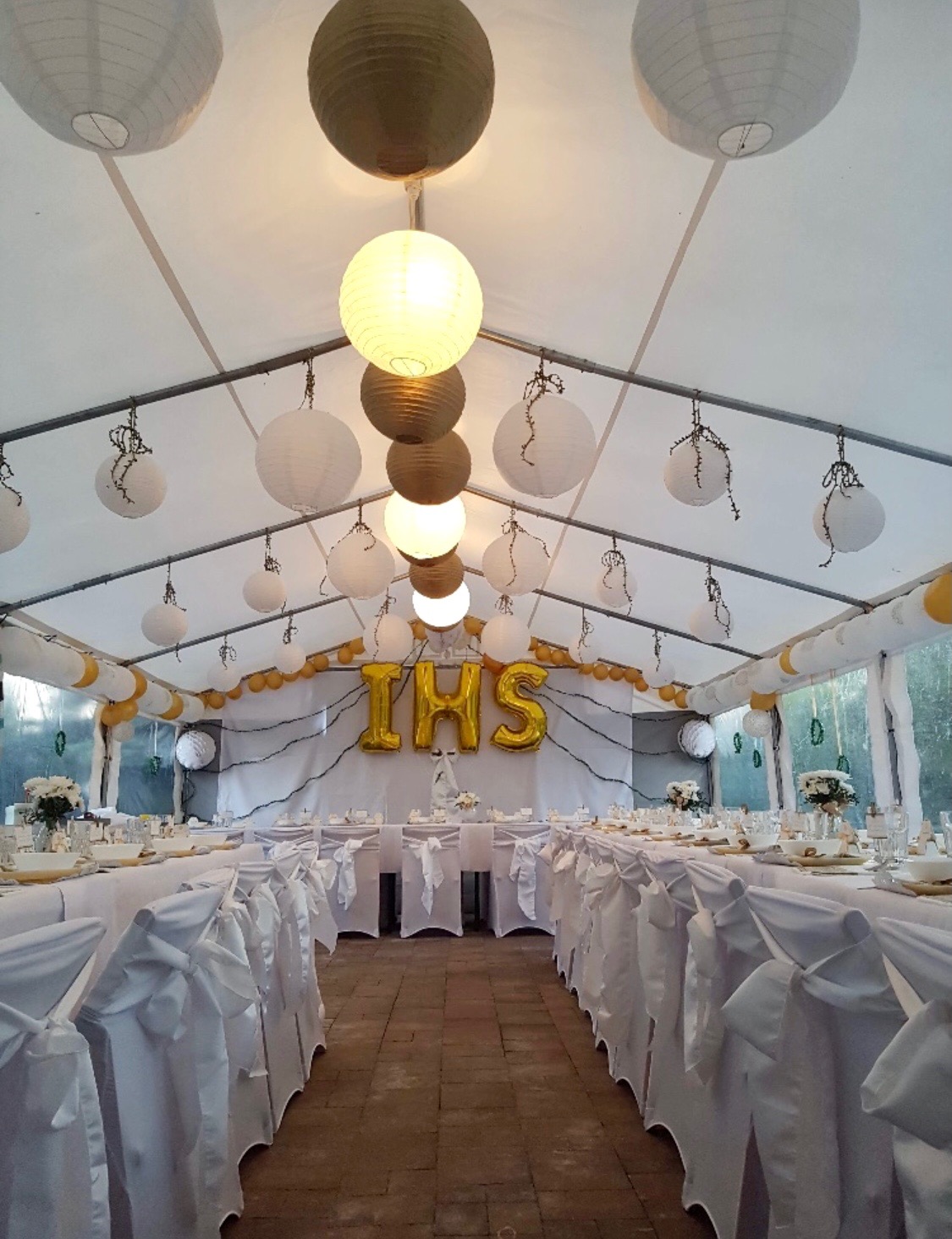 Uroczystość w białym namiocie: Długie stoły z białymi obrusami i krzesłami z dekoracyjnymi kokardami, oświetlone papierowymi lampionami i girlandami z balonów, z napisem IHS z balonów w tle.