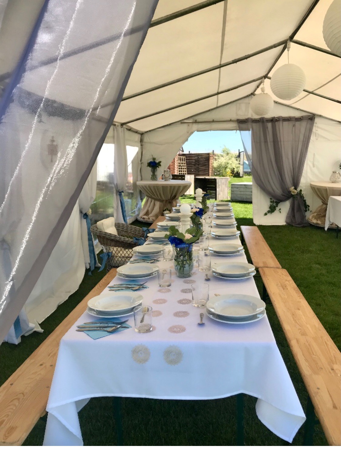 Uroczysty stół nakryty białym obrusem z dekoracyjnymi rozetami w namiocie, z drewnianymi ławkami po bokach, przygotowany na przyjęcie, widoczne dekoracje kwiatowe i wiszące lampiony.