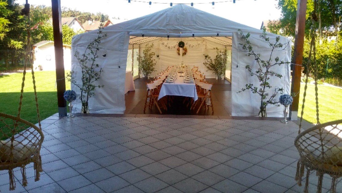 Ustawiony stół w białym namiocie imprezowym, udekorowanym kwiatami i girlandami świetlnymi, widok z zewnątrz z huśtawkami na pierwszym planie.