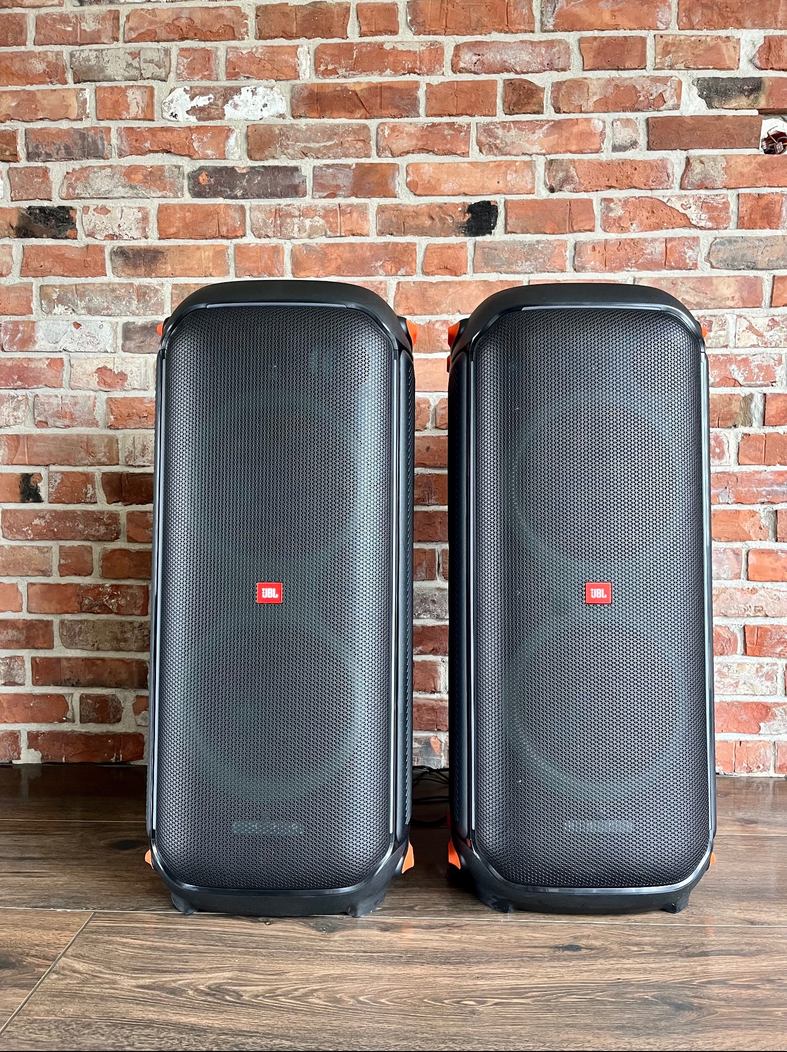 Dwa czarne głośniki estradowe JBL z pomarańczowymi akcentami, ustawione na drewnianej podłodze na tle ceglanej ściany.