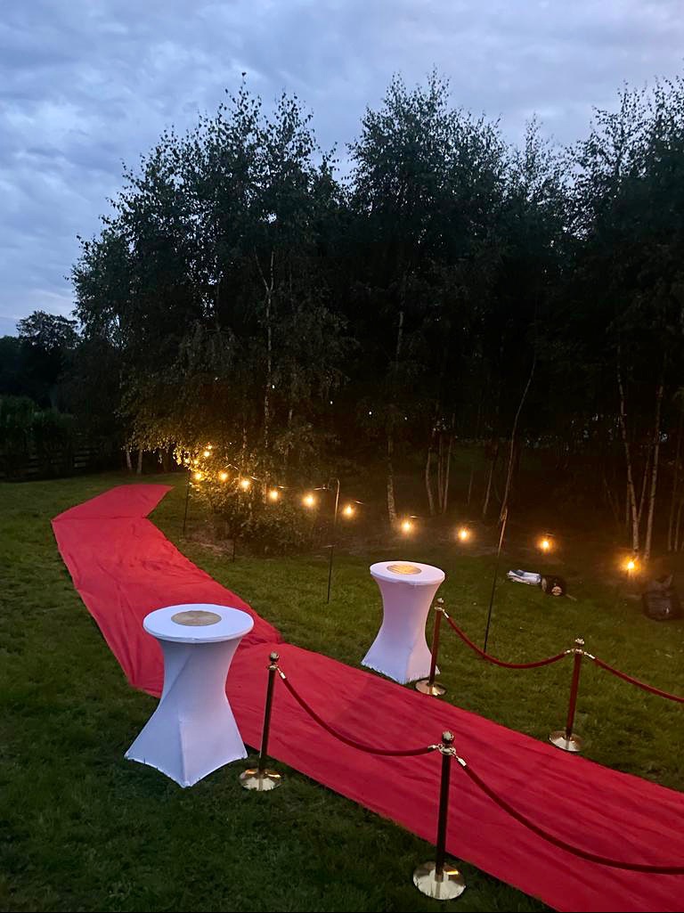 Czerwony dywan rozłożony na trawie, oświetlony girlandami żarówek, flankowany białymi stolikami koktajlowymi i złotymi słupkami z czerwonym sznurem, prowadzący w kierunku ciemnego lasu o zmierzchu.