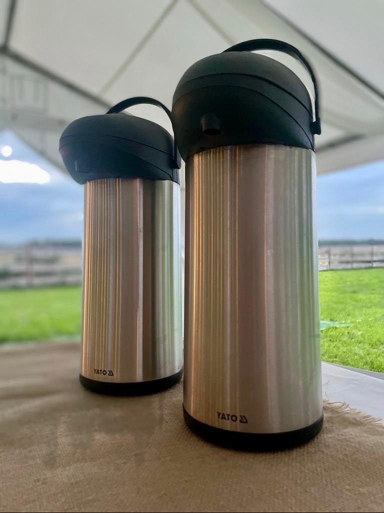 Dwa termosy marki YATO na stole, w tle widok na zieloną trawę i konstrukcję namiotu.
