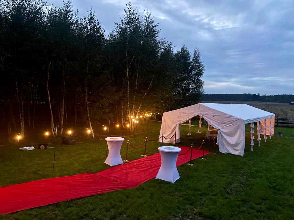 Biały namiot z czerwonym dywanem i dekoracyjnym oświetleniem w plenerze, przygotowany na imprezę wieczorową