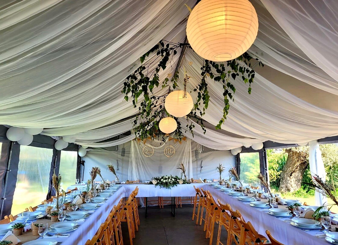 Udekorowany namiot weselny z białymi lampionami, zwisającymi zielonymi gałązkami i długim stołem z nakryciami, przygotowany na przyjęcie.