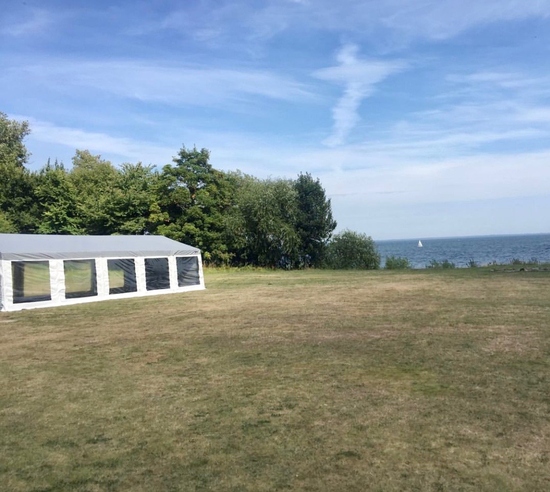 Szary namiot plenerowy z oknami na trawiastym terenie, w tle drzewa i morze z żaglówką pod błękitnym niebem z chmurami.