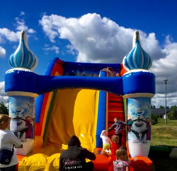 Kolorowy dmuchany zamek z motywem Frozen, dzieci bawiące się na zjeżdżalni i schodach, niebieskie niebo z chmurami w tle.