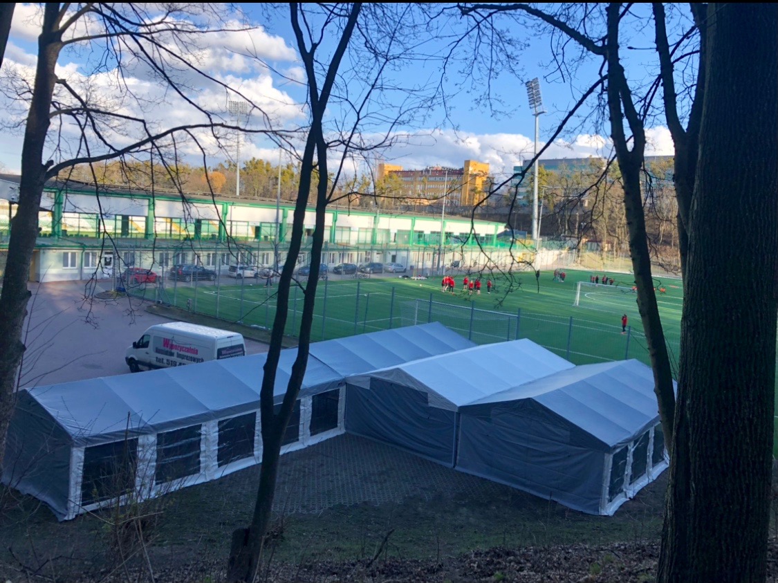Rozłożone szare namioty z oknami na tle boiska sportowego z trenującymi piłkarzami, widok z góry przez konary drzew. Na pierwszym planie fragmenty pni drzew.