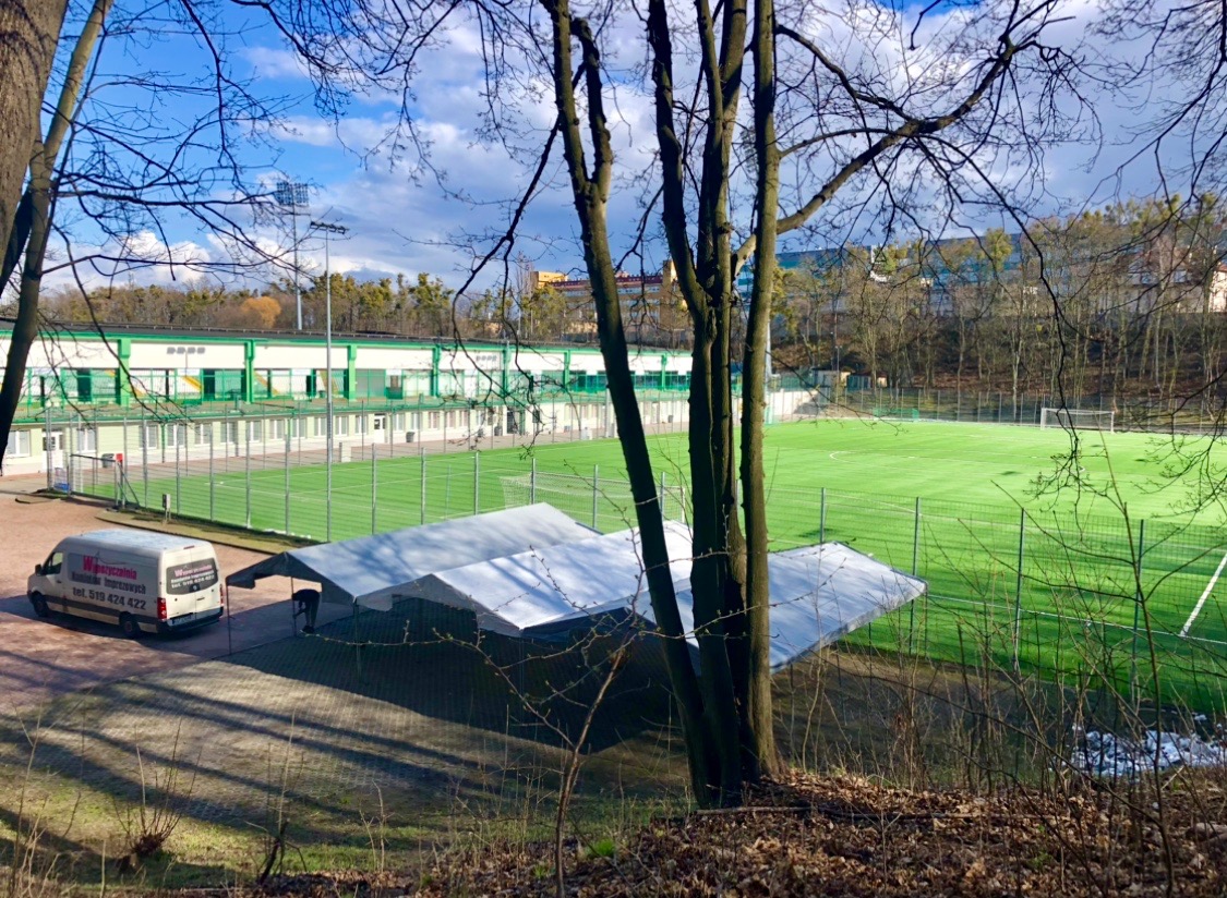 Rozłożony biały namiot plenerowy obok boiska piłkarskiego, widok zza drzew, na pierwszym planie uschnięte liście, w tle budynek z zielonymi akcentami i firmowy van z reklamą.