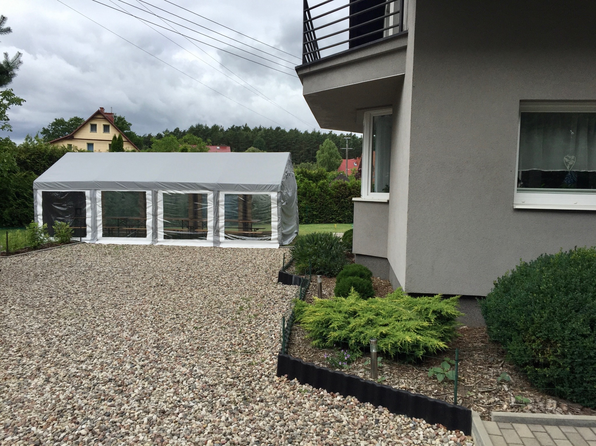Szary namiot plenerowy z przezroczystymi ścianami bocznymi, ustawiony na żwirowym podłożu obok nowoczesnego domu z szarym tynkiem i balkonem.