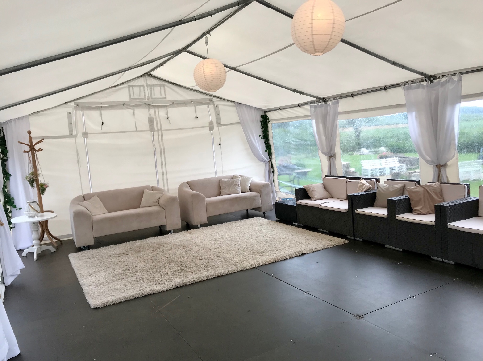 Wnętrze białego namiotu z beżowymi sofami, czarnymi fotelami rattanowymi, puszystym dywanem i papierowymi lampionami, dekoracja z zielonych pnączy przy oknach.