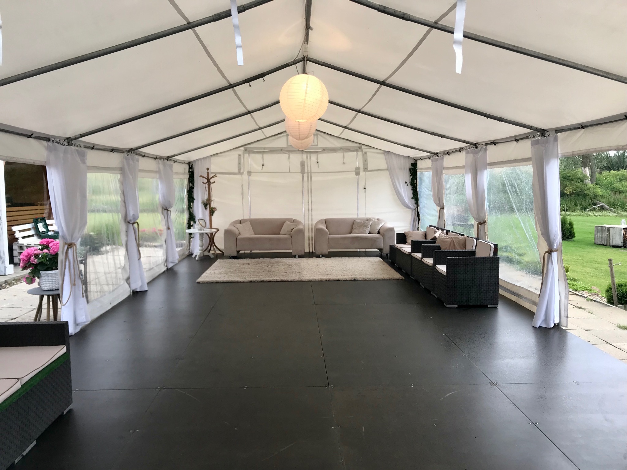 Wnętrze białego namiotu z czarną podłogą, urządzony w stylu lounge z beżowymi sofami, ciemnymi fotelami rattanowymi, białym dywanem i papierowymi lampionami wiszącymi u góry.
