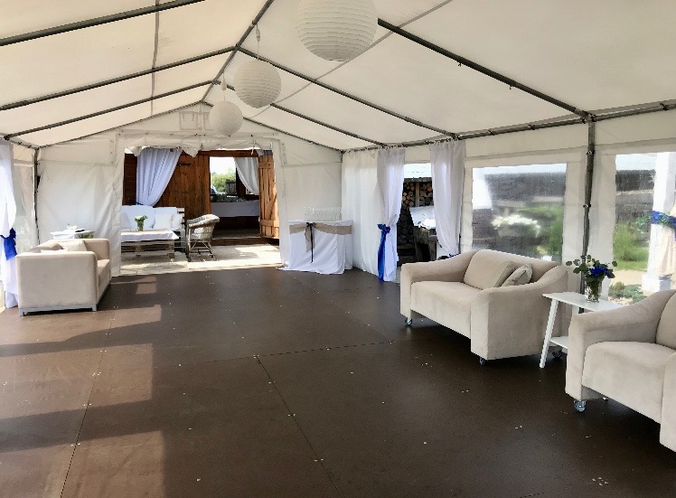 Wnętrze białego namiotu z drewnianą podłogą, wyposażonego w beżowe sofy, stoliki i dekoracje kwiatowe, oświetlone białymi lampionami.