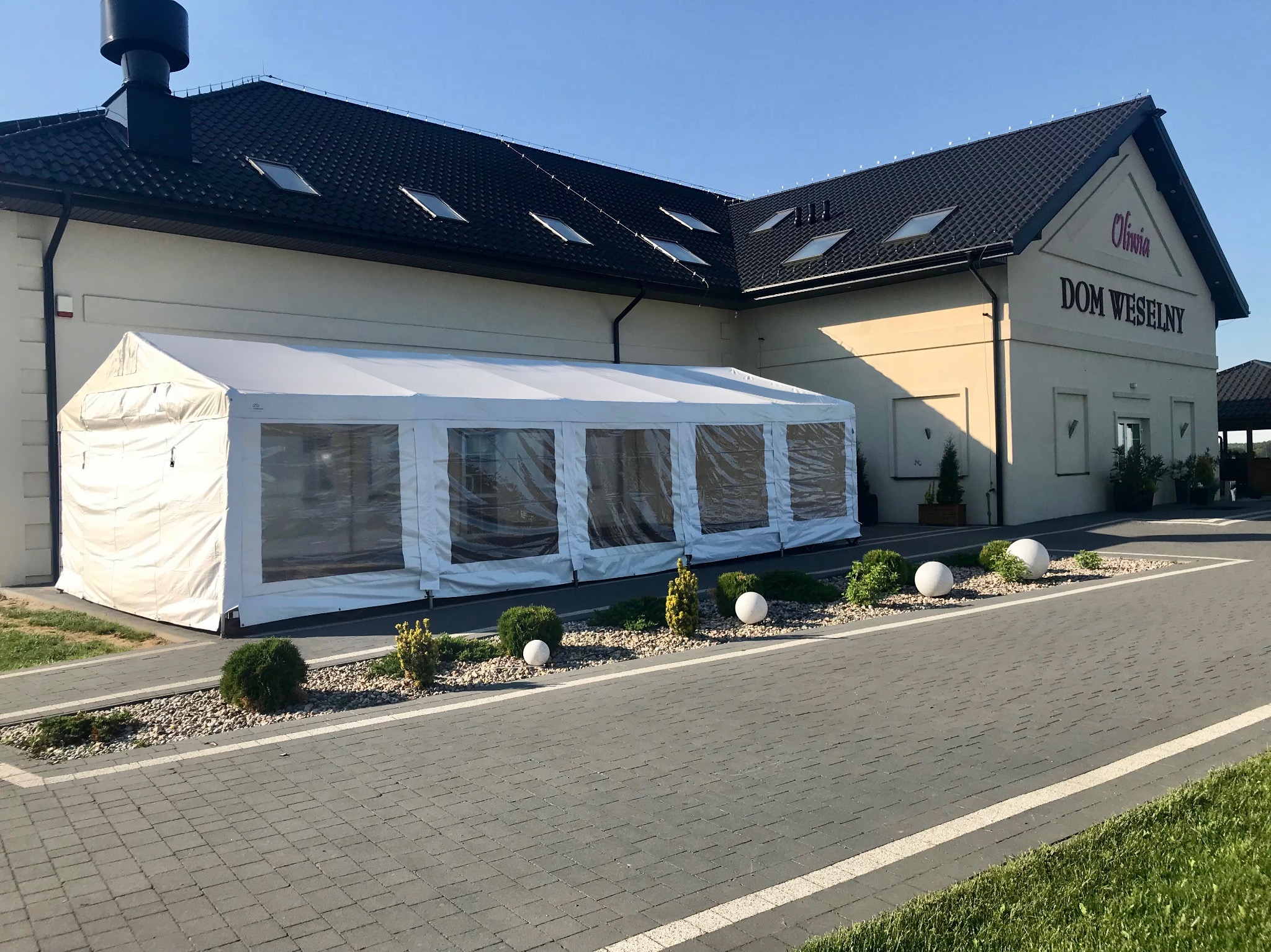 Biały namiot z przezroczystymi ścianami dostawiony do budynku Domu Weselnego Olivia, widok z zewnątrz, dekoracyjne kule i krzewy przed namiotem.