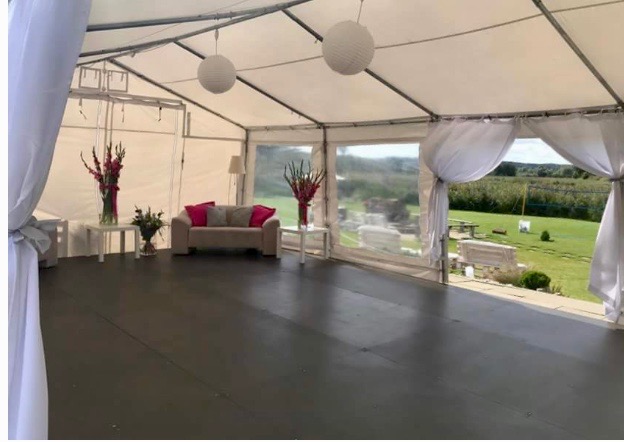 Wnętrze białego namiotu bankietowego z sofą, stolikami z kwiatami i papierowymi lampionami, widok na zielony teren przez okna.