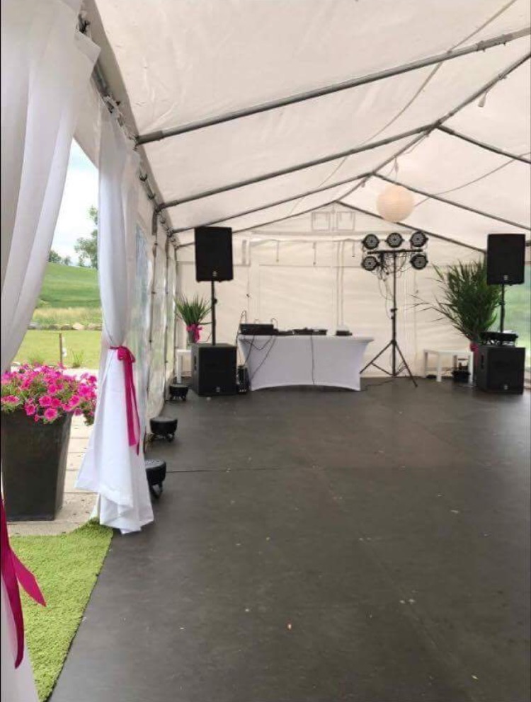 Wnętrze białego namiotu eventowego z nagłośnieniem, oświetleniem, stanowiskiem DJ-a i dekoracjami roślinnymi, widok na zielony teren na zewnątrz.