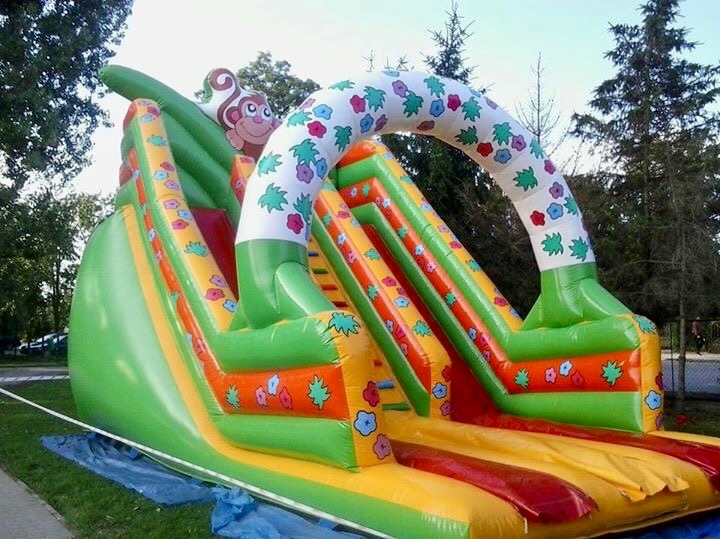 Kolorowa, dmuchana zjeżdżalnia z motywem małpki i kwiatów, rozłożona na trawie z widocznym niebieskim podkładem zabezpieczającym.