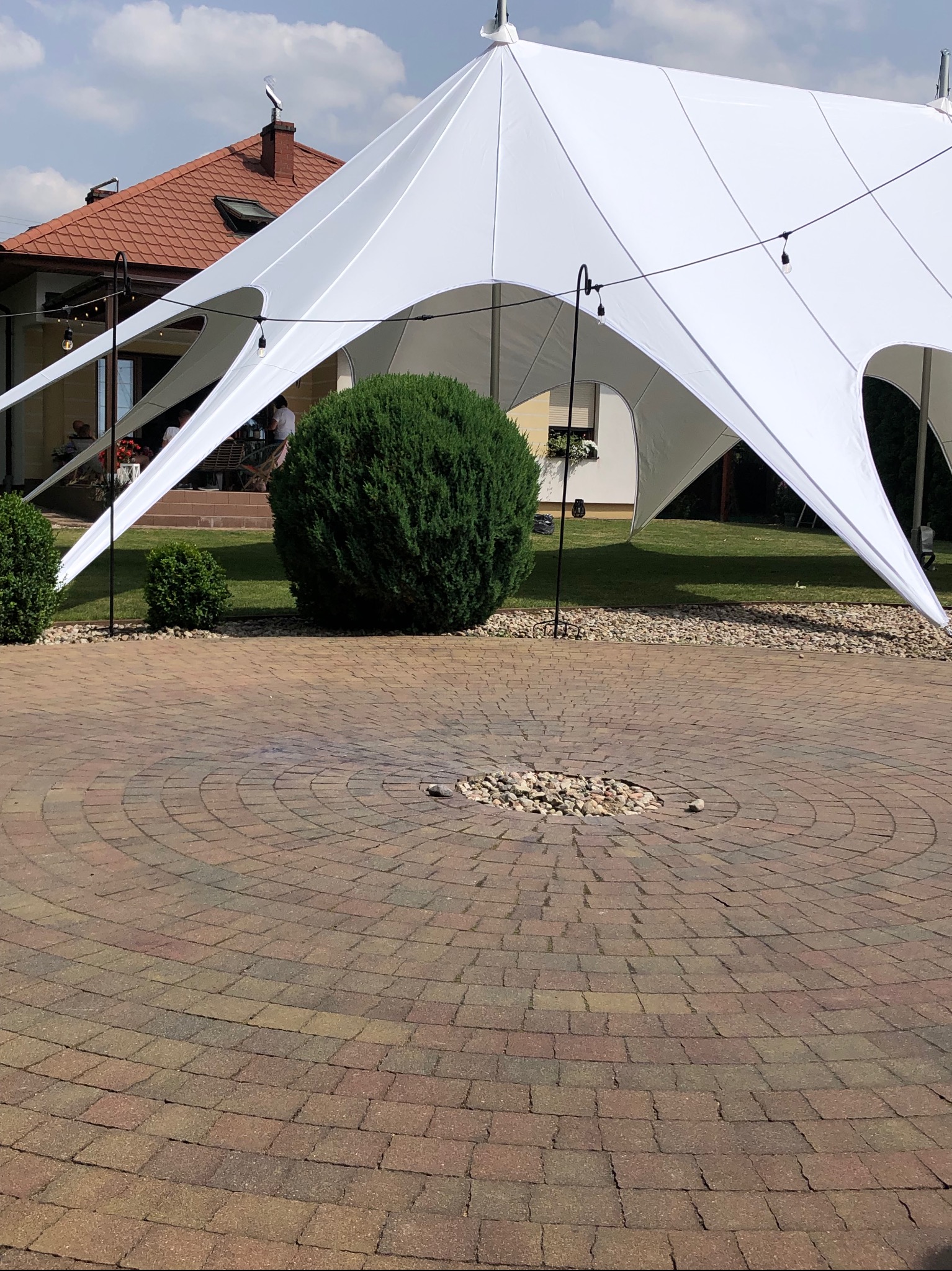 Biały namiot rozłożony na zewnątrz, widoczne okrągłe ułożenie kostki brukowej na ziemi i krzewy w tle, oświetlenie w postaci żarówek zwisających z namiotu.