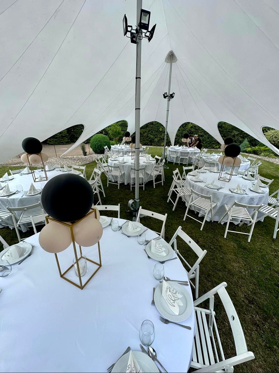Uroczyste stoły z białymi obrusami i dekoracjami balonowymi w czerni i beżu pod namiotem w kształcie gwiazdy, otoczone zielonym trawnikiem i krzewami.